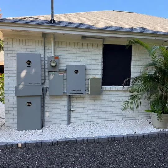 An EG4 FlexBOSS21 inverter mounted above its battery, the utility meter, an EG4 GridBOSS, and an Enphase IQ Combiner in one row on a white brick wall, with conduit running between them